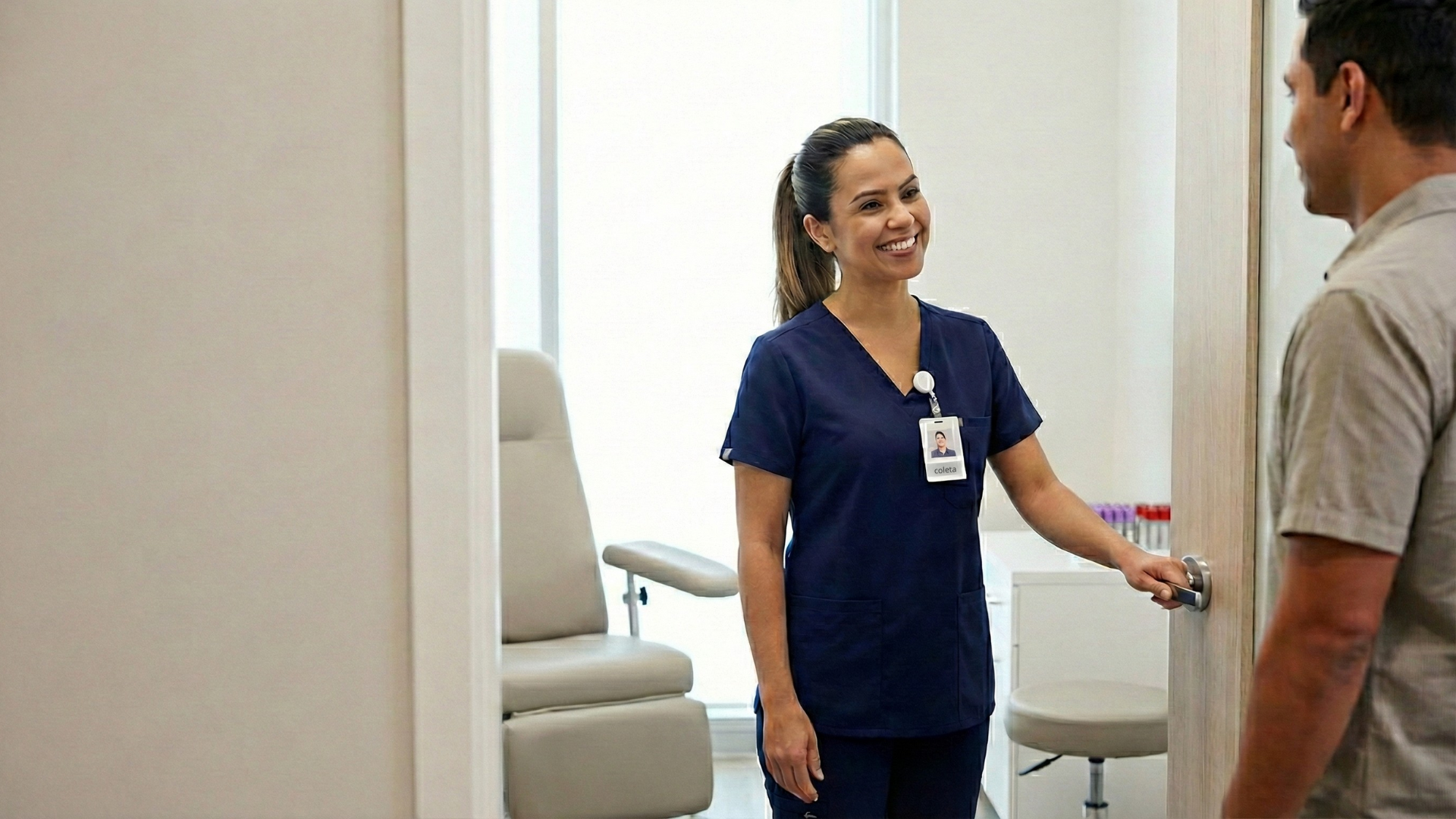 Profissional de coleta em uniforme azul marinho recebe paciente com sorriso na porta da sala de coleta de um laboratório de análises clínicas, transmitindo acolhimento, confiança e uma primeira impressão positiva durante a experiência do paciente.